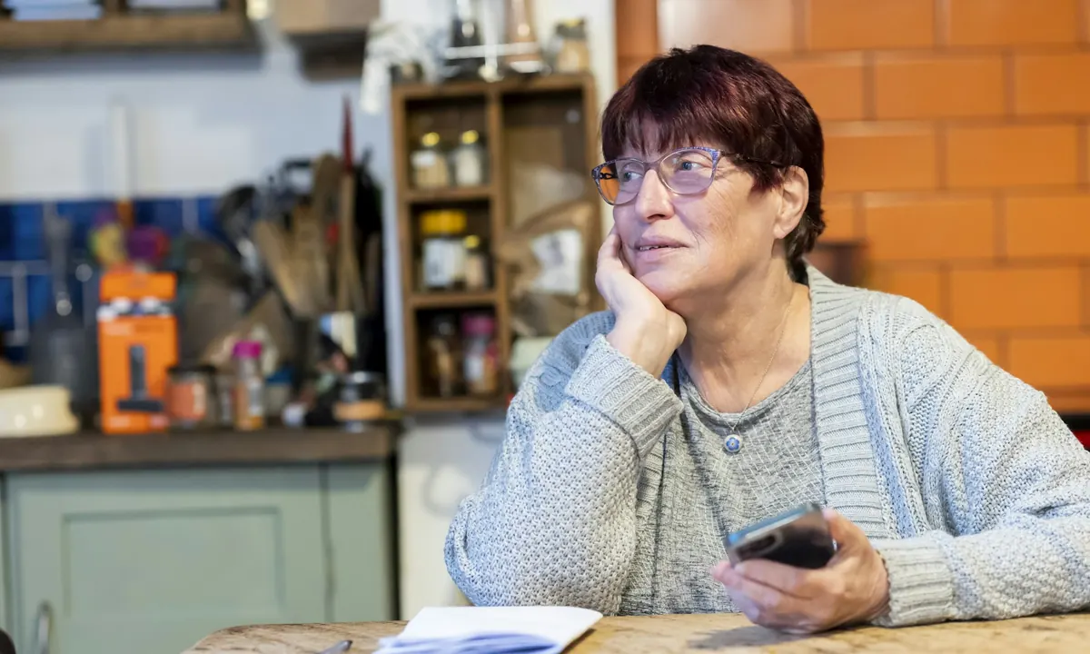 A woman at her kitchen table, thinking, with her phone in her hand